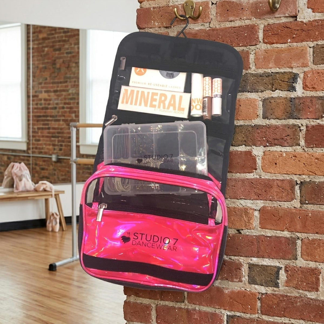 A black hanging makeup organizer pouch with a bright pink front pocket, shown open and filled with cosmetics and a clear organizer tray on a light gray background. The front pocket has the visible brand name STUDIO 7 DANCEWEAR printed on it.