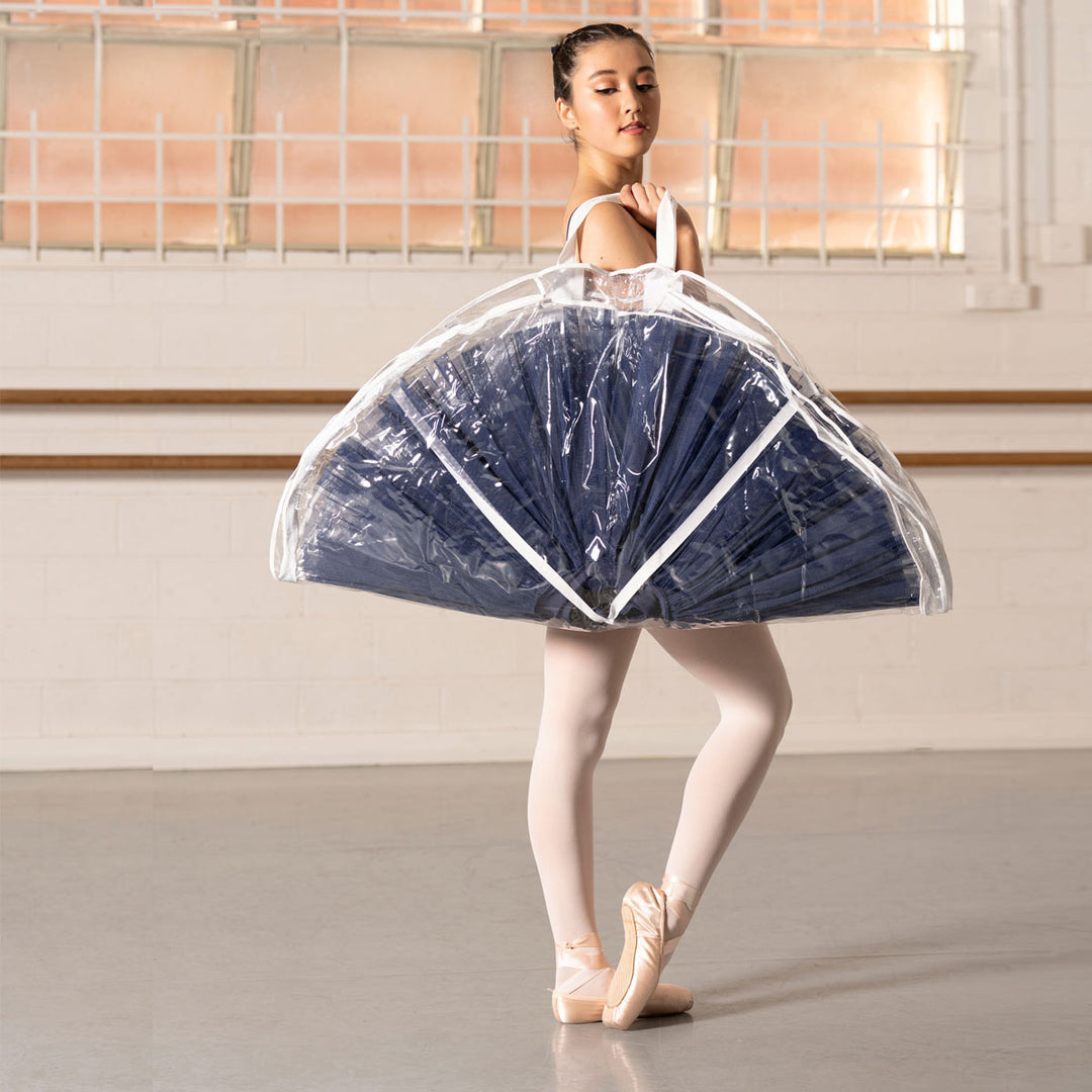 A ballerina in a navy blue tutu skirt and pointe shoes stands in a studio holding a large, clear plastic garment bag over her shoulder.