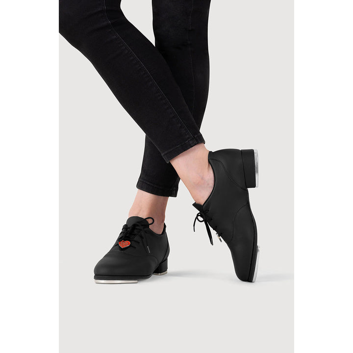 A person wearing black pointed-toe lace-up shoes with low block heels and black skinny jeans, shown from the calves down against a light gray studio background.
