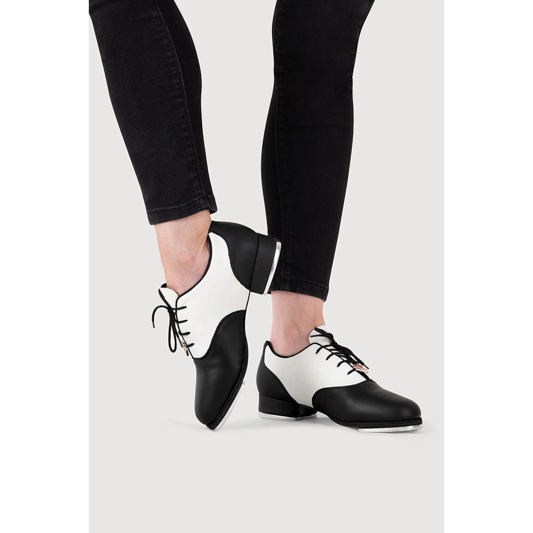 Black and white leather dance shoes with laces, shown on a model wearing black skinny jeans against a light gray background.