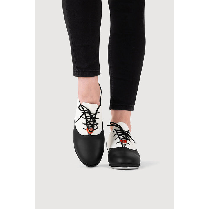 A person wearing black and white leather lace-up shoes with black laces and a small orange accent near the laces, paired with black skinny jeans, shown against a light gray background.