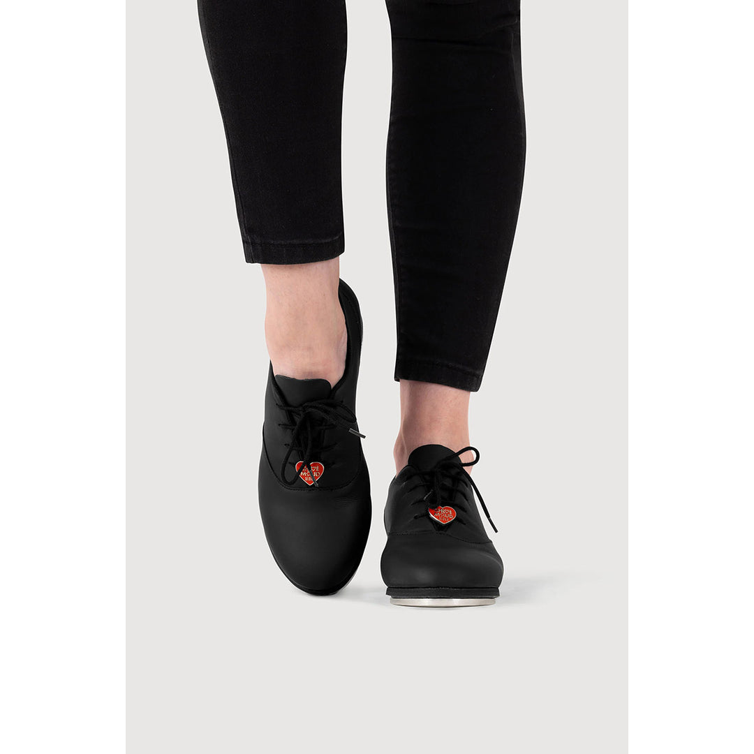 Black lace-up shoes with small red heart details on the sides, shown on a model wearing black pants against a light gray background.