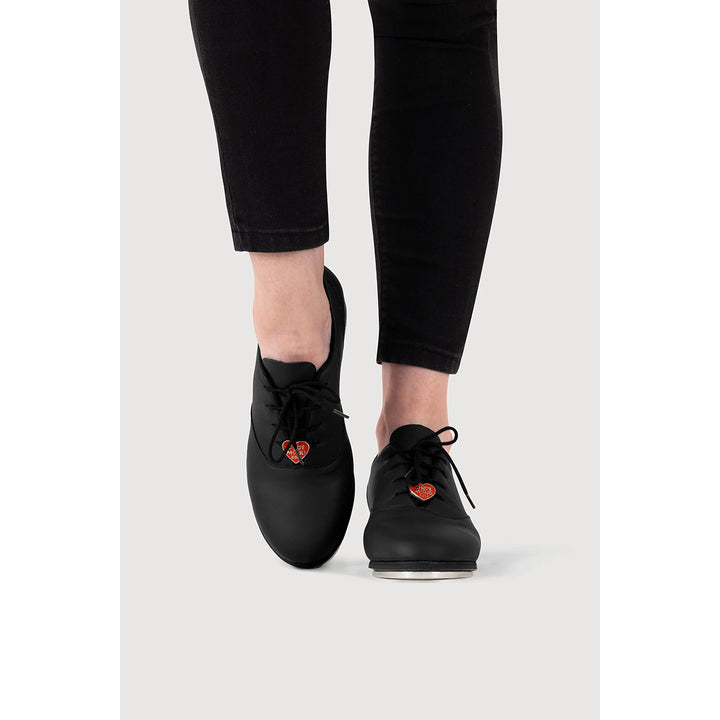 Black lace-up shoes with small red heart details on the sides, shown on a model wearing black pants against a light gray background.