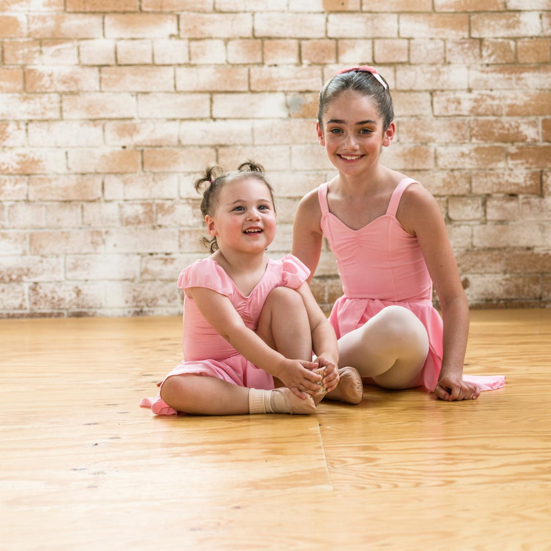 Two young girls in pink ballet leotards sitting on a wooden floor in front of a brick wall.